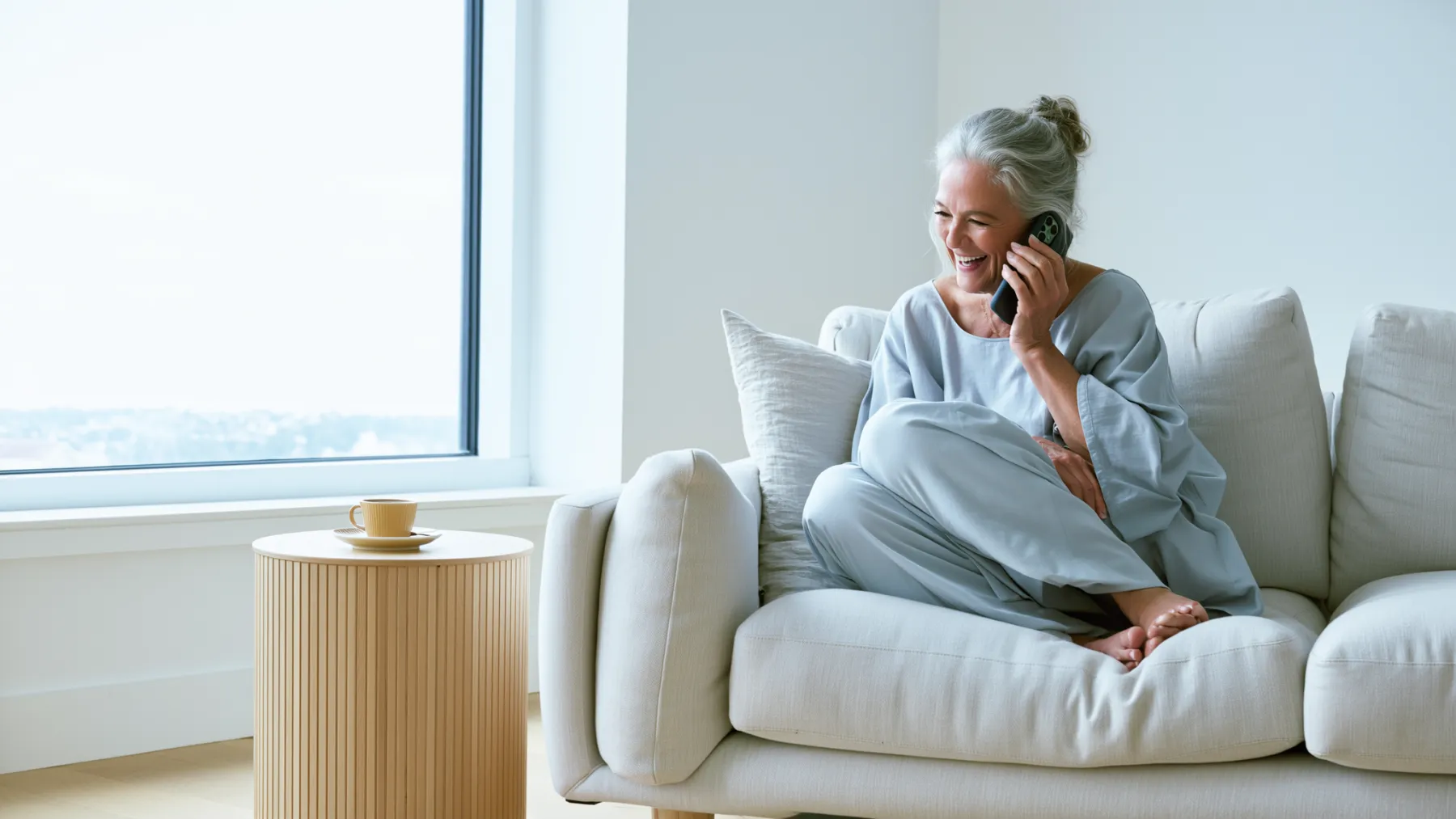 A grandmother laughing on the phone on her sofa by a large window